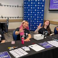 Four students sitting at registration table giving anchor up sign